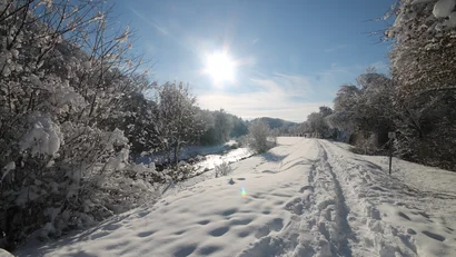 Verschneiter Weg neben Fluss im Wald bei Sonnenschein